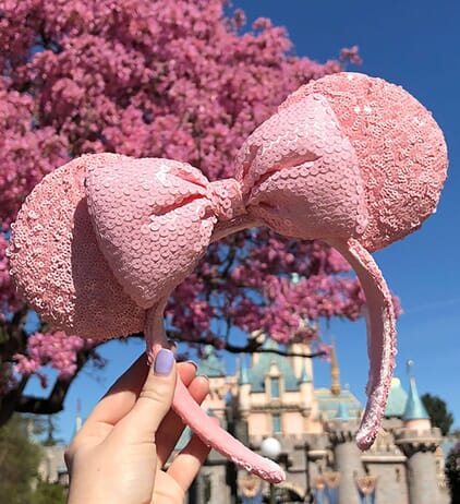 Woman's hand holding pink sequined Minnie Mouse ears in front of Disneyland castle