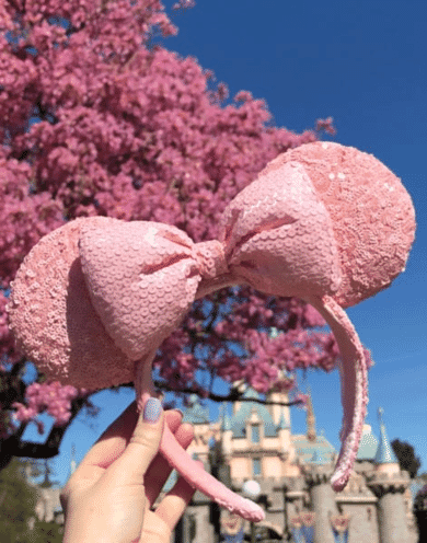 Woman's hand holding pink sequined Minnie Mouse ears in front of Disneyland castle