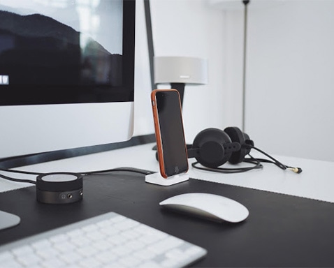 iPhone in orange case on white stand on top of white desk next to white mouse, keyboard and monitor