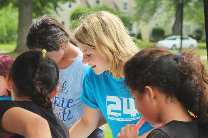 Woman in blue t-shirt talking with a group of children on a green grassy lawn