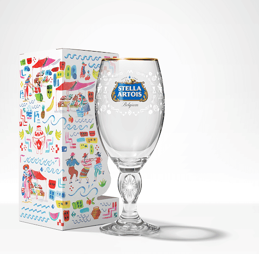 Clear glass Stella Artois-branded chalice with gold rim and blue logo next to colorful gift box