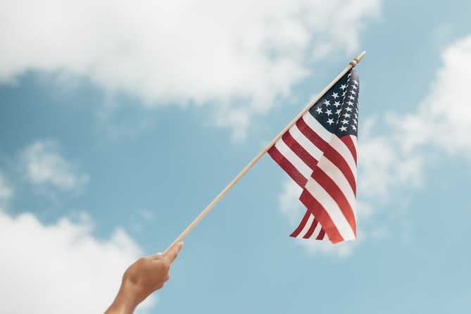 Person's hand waving an American flag in front of a blue sky and white clouds.