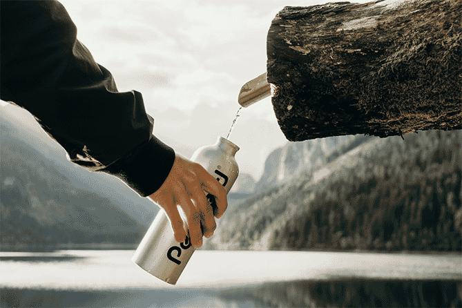 Man holding stainless steel tumbler with black logo in front of a lake and mountains
